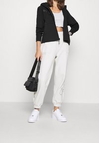 Black zip-up hoodie with hood, white cropped top, cream joggers with pockets and logo, black shoulder bag, white sneakers.