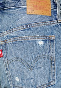 Světle modré džíny z denimu s opotřebovaným zadním kapsou, žlutým prošíváním a hnědou koženou etiketu s informacemi o značce a modelu.