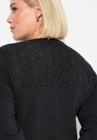 Camiseta negra con un panel de encaje floral bordado texturizado a lo largo de los hombros. Mangas largas y cuerpo de tela suave.