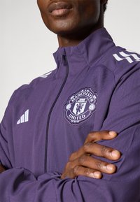 Lila Sportjacke mit Reißverschluss vorne, Adidas drei Streifen auf den Schultern und gesticktem Manchester United Logo auf der Brust.