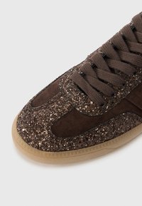Sneaker marrone con una tomaia glitterata, accenti in suede, punta rotonda e lacci piatti. La suola è in gomma traslucida con dettagli testurizzati.