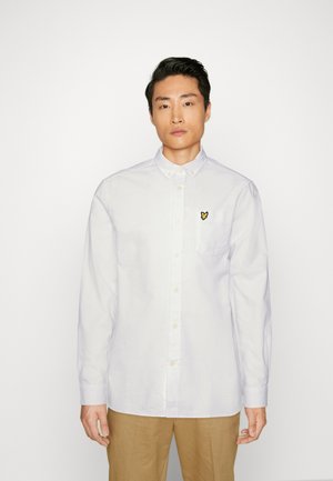Chemise blanche à boutons avec manches longues, présentant un petit logo brodé noir et jaune sur la poche gauche de la poitrine. Texture lisse.