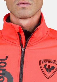 Ljus orange jacka med hög krage, svart dragkedja och en svart logotyp-etikett. Har ett mjukt, elastiskt material med synlig sömnad.