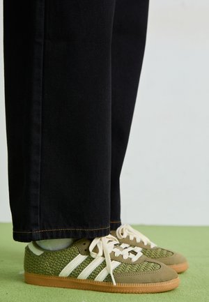 Zielone plecione trampki z białymi paskami i gumowymi podeszwami, noszone z czarnymi spodniami na jasnozielonej podłodze.