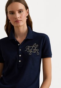 Tumesinise polo särk, millel on tekstuuriga hõbedane logo, klassikalise krae ja kolme nupuga avamine. Pehme puuvillane materjal.