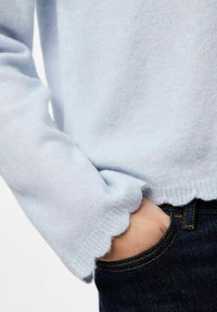 Ljusblå tröja med en vågig nederkant, gjord av mjukt material, matchad med mörk denimjeans. Hand placerad i framfickan.