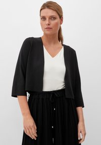 Zwarte cropped cardigan met driekwartmouwen, gedragen over een witte V-hals top. Het model combineert het met een geplooide zwarte rok. Glad textuur.