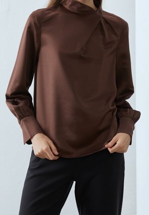 Brown, satin blouse with a high neckline, long sleeves, and gathering details at the collar. Paired with black pants.
