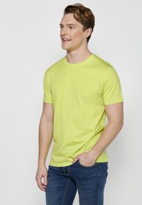 Tricou scurt cu mâneci verzi lime, realizat dintr-un material neted, având un logo subtil pe piept și un guler clasic tip crew. Asortat cu blugi albaștri.