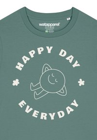 T-shirt en coton vert avec un texte circulaire blanc "JOUR HEUREUX TOUS LES JOURS" et un graphique de soleil souriant, orné de petites fleurs.