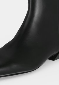 Bottine noire en cuir lisse avec un bout pointu et un talon bas incliné sur un fond blanc.