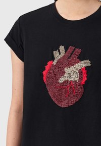 Κοντινό πλάνο ενός μαύρου T-shirt με λεπτομερές σχέδιο ανατομικής καρδιάς από χάντρες, σε κόκκινο και μπεζ, στο ύψος του στήθους.