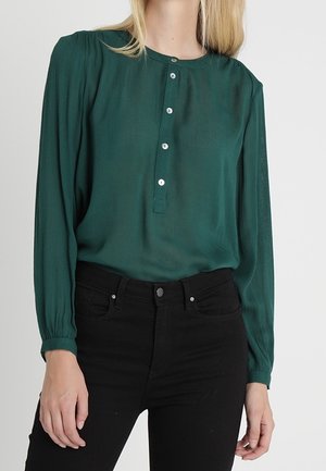 Kobieta ubrana w ciemnozieloną bluzkę z długim rękawem i guzikami z przodu, wpuszczoną do czarnych spodni z wysokim stanem.