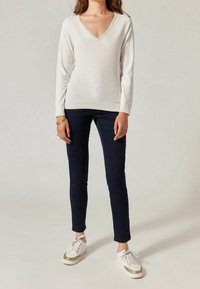 Kobieta w kremowym swetrze z długim rękawem i dekoltem w serek, ciemnych obcisłych spodniach oraz białych i złotych sneakersach, stojąca na tle jednolitego jasnego tła.