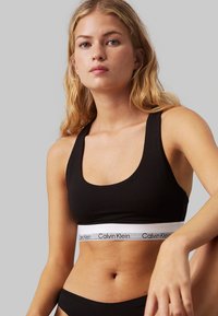 Sujetador deportivo negro con una amplia banda blanca que presenta un logo negro de "Calvin Klein". Textura suave, escote redondo y tirantes anchos.