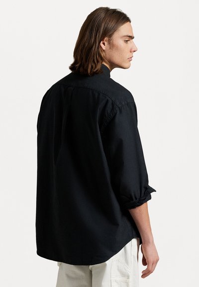 Camiseta negra de manga larga con un corte relajado, cuello botón y puños enrollados. Textura suave y dobladillo ligeramente curvado.