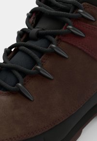 Brown suede hiking shoe featuring black fabric accents, textured laces, and cone-shaped black lace tips. Durable sole designed for rugged terrain.