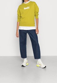 Sudadera verde con el logo blanco de Levi's, superpuesta sobre una camisa blanca, combinada con jeans de pierna ancha azul oscuro y zapatillas blancas con acentos neón.