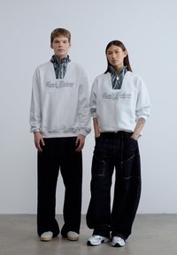 Λευκό φούτερ με φερμουάρ στο γιακά και λογότυπο, συνδυασμένο με άνετο, σκουρόχρωμο τζιν παντελόνι. Τα μοντέλα φορούν λευκά και μαύρα παπούτσια. Ουδέτερο φόντο.