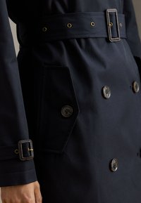 Trench-coat bleu marine avec taille ceinturée, boutons apparents et tissu texturé. Comprend deux poches avant et des accents dorés.