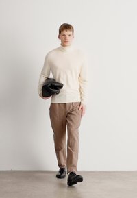 Pulover bej cu guler înalt, tricot ușor; pantaloni bej deschis, croi drept; geanta neagră strânsă în mâna dreaptă; pantofi negri.