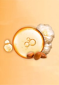 Gyllene olja i en rund skål omgiven av två mindre droppar, bruna argannötter och en vit blomma mot en orange bakgrund.
