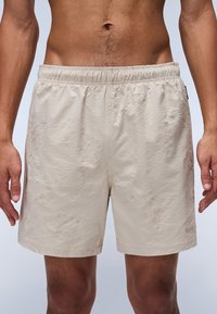 Beige zwemshorts gemaakt van een textuurstof, met een elastische tailleband en subtiele abstracte patronen overal. Geen zichtbare zakken.