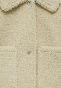 Veste en tissu sherpa couleur crème à l'avant avec deux poches, fermeture à boutons-pression et col large, présentant un matériau texturé semblable à de la laine.