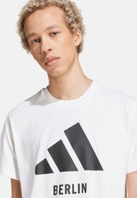 Witte katoenen t-shirt met een grote zwarte afdruk van drie diagonale strepen en het woord "BERLIJN" eronder gedrukt. Zachte textuur, ontspannen pasvorm.