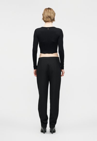 Femme aux cheveux blonds courts portant un crop top noir ajusté à manches longues, un pantalon noir taille haute et des chaussures à talons noires, vue de dos.