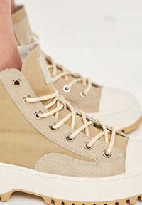 Béžové vysoké plátěné a semišové tenisky s tlustými bílými gumovými podrážkami a béžovými tkaničkami, které jsou obuty na nohách na světlém pozadí.