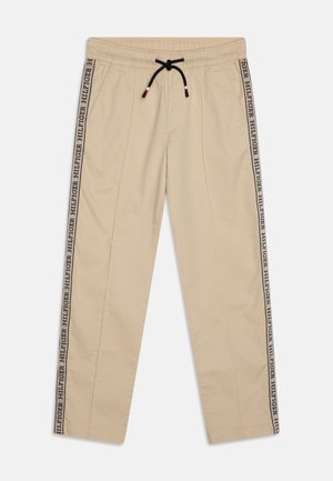 Pantalons beige à coupe décontractée avec une taille élastique et un cordon de serrage, arborant le logo noir "HILFIGER" sur les côtés. Texture de tissu lisse.