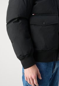Jachetă puffer neagră cu o finisare fină, manșete elastice, buzunar lateral și detalii subtile cu logo. Purtați-o peste blugi negri.