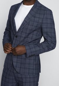 Marinblå rutig kavaj med notch laplar, en knappstängning och detaljer med fyraknappsmuddar. Bärs över en vit t-shirt.