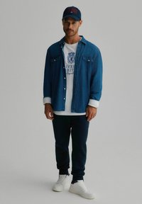 Koszula dżinsowa z dwoma kieszeniami na piersi, biała koszulka z niebieskim logo, granatowe dresy i białe sneakersy. Casualowy strój z nałożonymi warstwami.