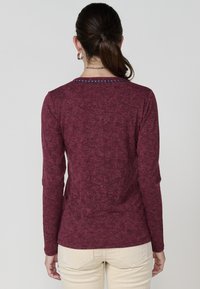 Ilgi piedurkņu, bordo krāsas tops ar teksturētu dizainu un pērlītēm rotātu apkakles detaļu. Mīksts audums, pieguļošs siluets un klasiskā apaļā kakla stila. 