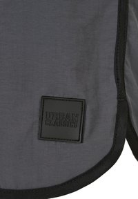 Un tissu gris avec une texture lisse présente un patch noir proéminent portant l'inscription "URBAN CLASSICS" en lettres en relief. Un contour noir souligne les bords.