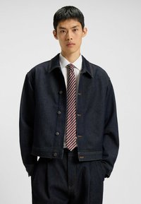 Mørk denimjakke med en boxy pasform, der har fem knaplukninger, kontrasterende syninger og sidelommer, parret med en hvid skjorte og et stribet slips.