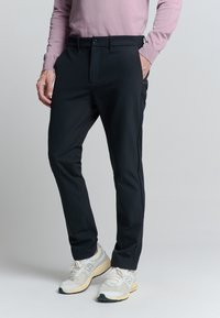 Man in een zwarte slim-fit broek, lichtroze langarmshirt en witte hardloopschoenen, staand met één hand in de zak.
