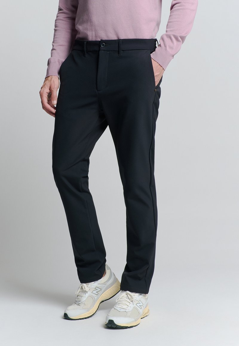 Man in een zwarte slim-fit broek, lichtroze langarmshirt en witte hardloopschoenen, staand met één hand in de zak.