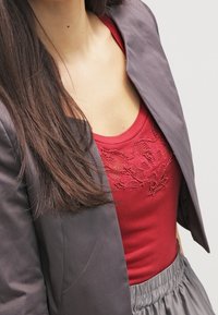 Femme aux longs cheveux bruns portant un blazer gris foncé sur un haut rouge brodé et un pantalon gris taille haute, torse visible.