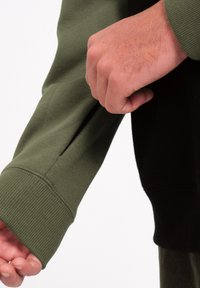 Sweat-shirt vert avec un poignet côtelé, présentant une poche zippée sur la partie supérieure de la manche. Le tissu a une texture lisse avec des détails noirs contrastants.