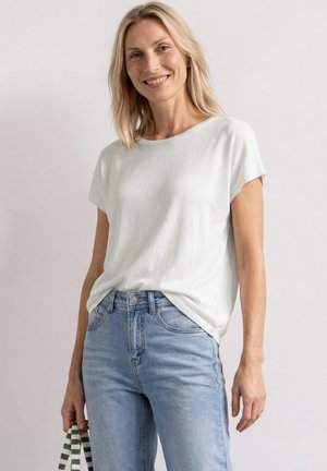 Smilende kvinde med blondt hår iført en hvid kortærmet top og lyseblå jeans, der holder en grøn og hvid stribet taske.