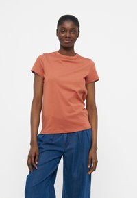 Γυναίκα που στέκεται και χαμογελά, φορώντας ένα απλό κοντομάνικο μπλουζάκι σε χρώμα σκουριάς και άνετο μπλε τζιν παντελόνι, μπροστά σε λευκό φόντο.