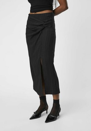 Jupe noire midi avec des rayures verticales texturées, présentant un détail avant torsadé et une fente latérale. Associée à des escarpins noirs à bout pointu.