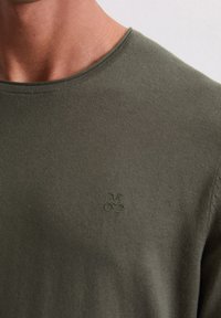 Olivgrünes Rundhals-T-Shirt aus weichem Stoff, mit einem dezenten bestickten Logo auf der Brust. Glatte Textur und figurnahes Design.