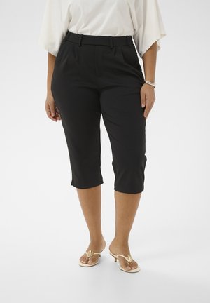 Femeie purtând pantaloni negri scurtați, bluză albă și sandale bej cu toc, stând pe un fundal alb.