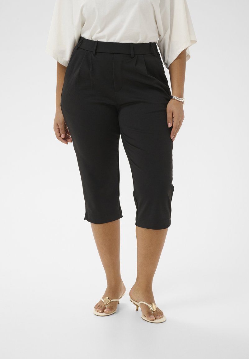 Femme portant un pantalon court noir, un chemisier blanc et des sandales à talons beige, debout sur un fond blanc.
