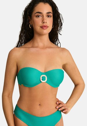 Vrouw met lang gekruld donker haar die een turquoise strapless bikini met een decoratieve gesp aan de bovenkant draagt, staand met hand op heup tegen een witte achtergrond.