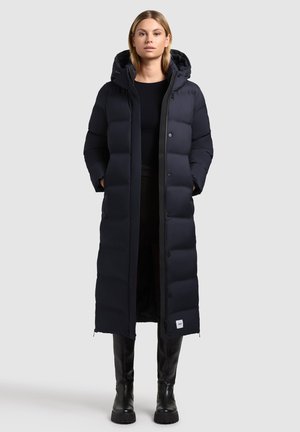 Lange pufferjas in donkerblauw, met een capuchon, gewatteerde buitenkant, drukknoopsluitingen en een merklabel nabij de zoom. Gemaakt van geïsoleerd materiaal.